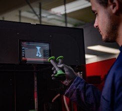 Una pieza rígida y flexible de múltiples materiales impresa con una impresora VLM, como se muestra detrás.